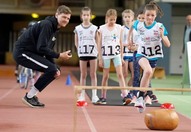 Kinder mit Startnummern auf Trikots in einer Reihe, ein Mädchen nimmt Anlauf, links daneben ein Mann gehockt mit Stoppuhr, im unscharfen Vordergrund Medizinball und Bank als Hindernisse